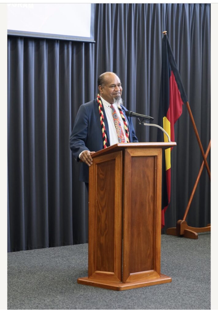 Vaughn McGuire speaking at a podium beside the Aboriginal flag